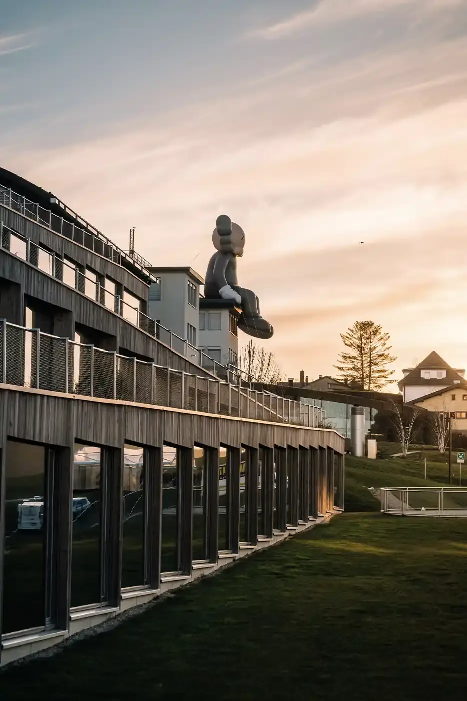 AllRightsReserved A massive 11-meter tall KAWS COMPANION sculpture in grey-toned color sits on the rooftop of the Audemars Piguet headquarters. The sculpture overlooks the glass spiral museum building in Le Brassus, Switzerland, against a mountainous backdrop, symbolizing the intersection of art and traditional watchmaking craftsmanship. (image 5 of 9)
