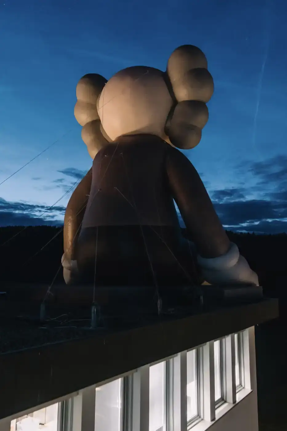 AllRightsReserved A massive 11-meter tall KAWS COMPANION sculpture in grey-toned color sits on the rooftop of the Audemars Piguet headquarters. The sculpture overlooks the glass spiral museum building in Le Brassus, Switzerland, against a mountainous backdrop, symbolizing the intersection of art and traditional watchmaking craftsmanship. (image 3 of 9)