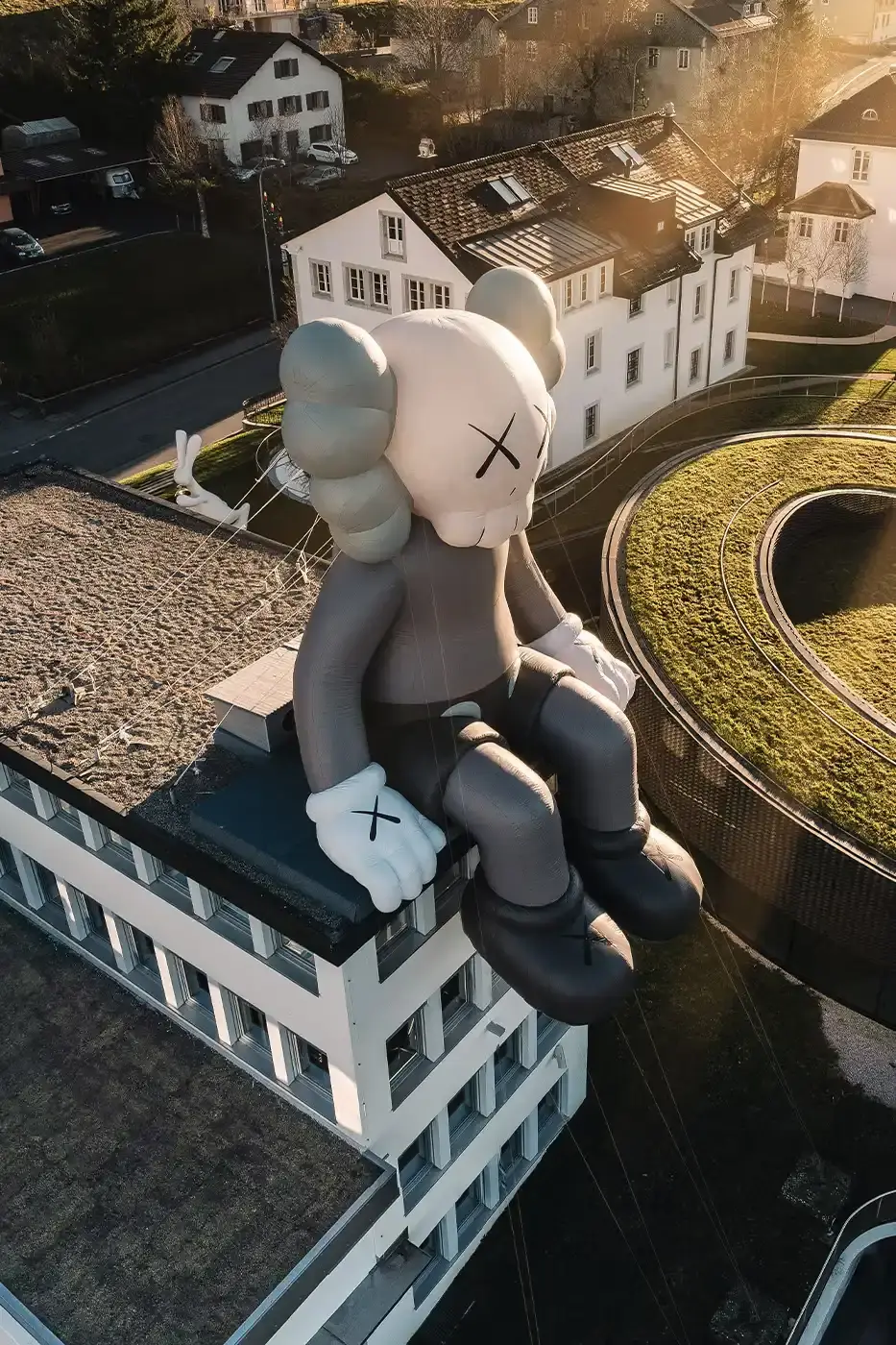 AllRightsReserved A massive 11-meter tall KAWS COMPANION sculpture in grey-toned color sits on the rooftop of the Audemars Piguet headquarters. The sculpture overlooks the glass spiral museum building in Le Brassus, Switzerland, against a mountainous backdrop, symbolizing the intersection of art and traditional watchmaking craftsmanship.