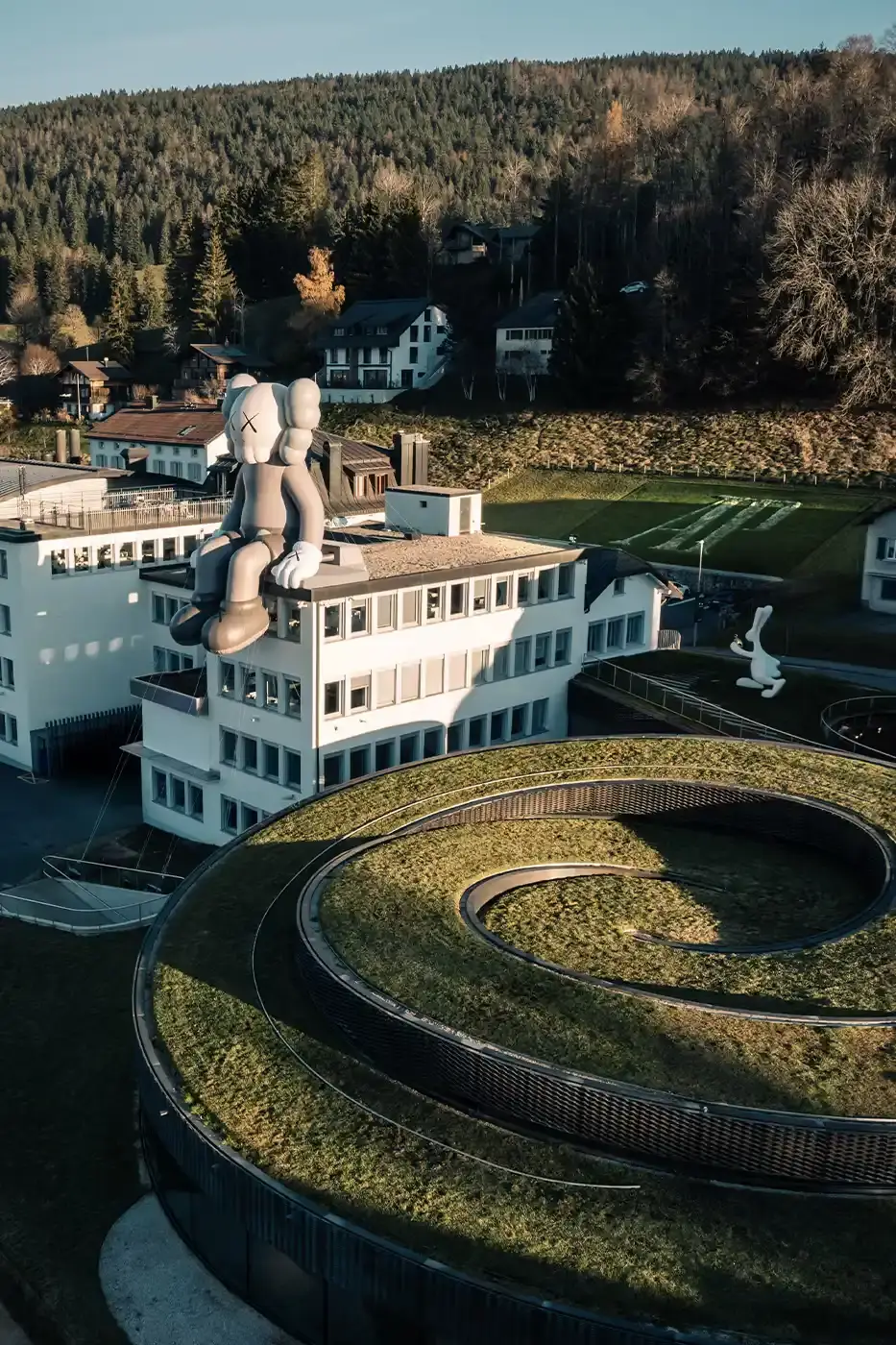 AllRightsReserved A massive 11-meter tall KAWS COMPANION sculpture in grey-toned color sits on the rooftop of the Audemars Piguet headquarters. The sculpture overlooks the glass spiral museum building in Le Brassus, Switzerland, against a mountainous backdrop, symbolizing the intersection of art and traditional watchmaking craftsmanship. (image 8 of 9)