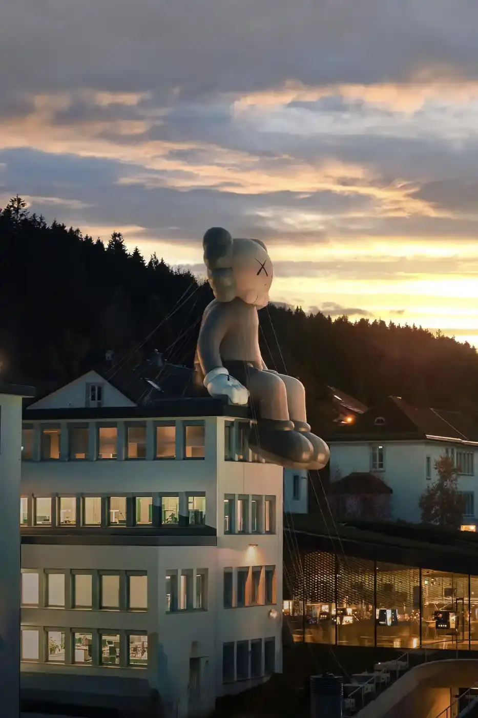 AllRightsReserved A massive 11-meter tall KAWS COMPANION sculpture in grey-toned color sits on the rooftop of the Audemars Piguet headquarters. The sculpture overlooks the glass spiral museum building in Le Brassus, Switzerland, against a mountainous backdrop, symbolizing the intersection of art and traditional watchmaking craftsmanship. (image 7 of 9)
