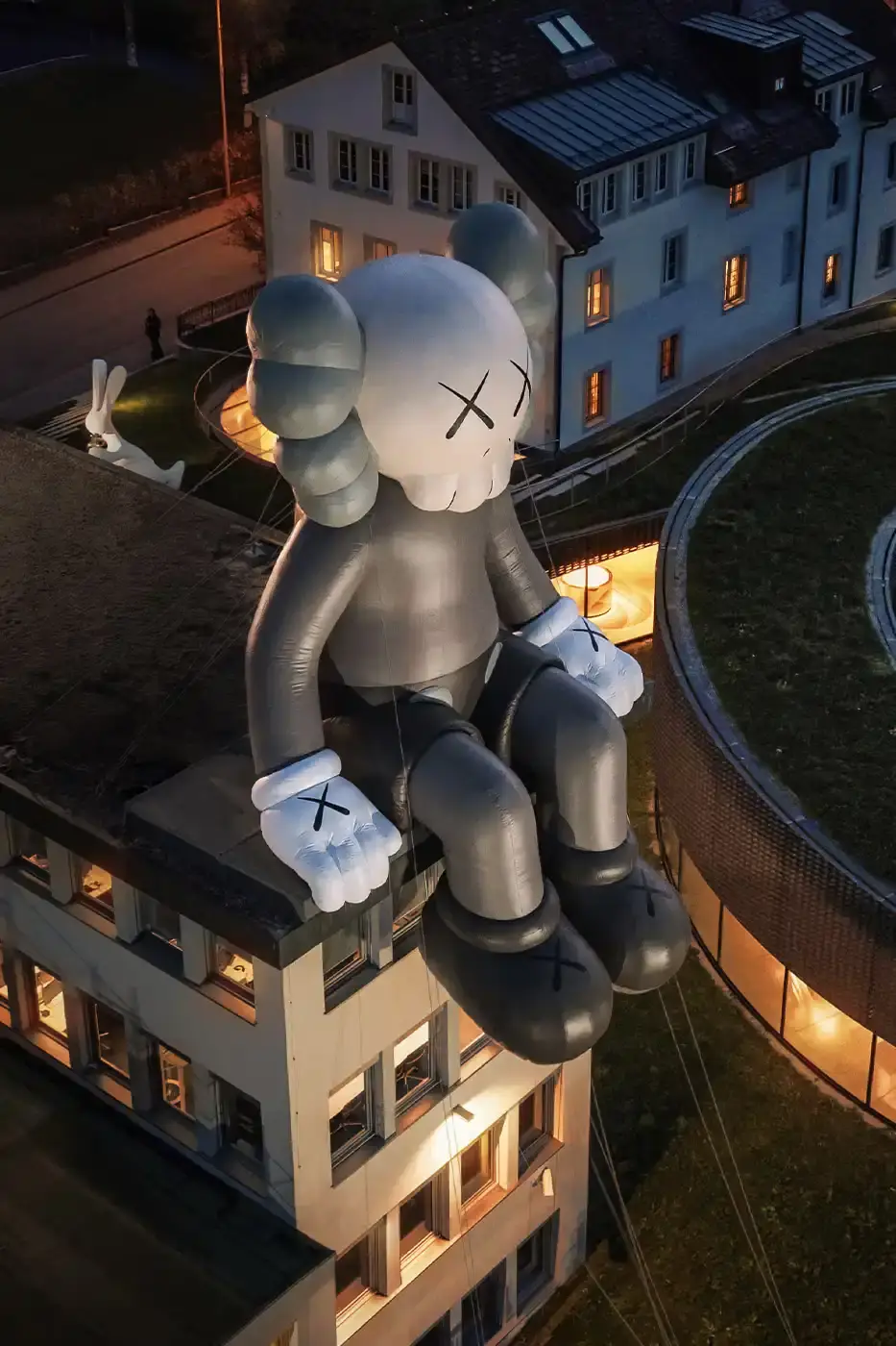 AllRightsReserved A massive 11-meter tall KAWS COMPANION sculpture in grey-toned color sits on the rooftop of the Audemars Piguet headquarters. The sculpture overlooks the glass spiral museum building in Le Brassus, Switzerland, against a mountainous backdrop, symbolizing the intersection of art and traditional watchmaking craftsmanship. (image 4 of 9)