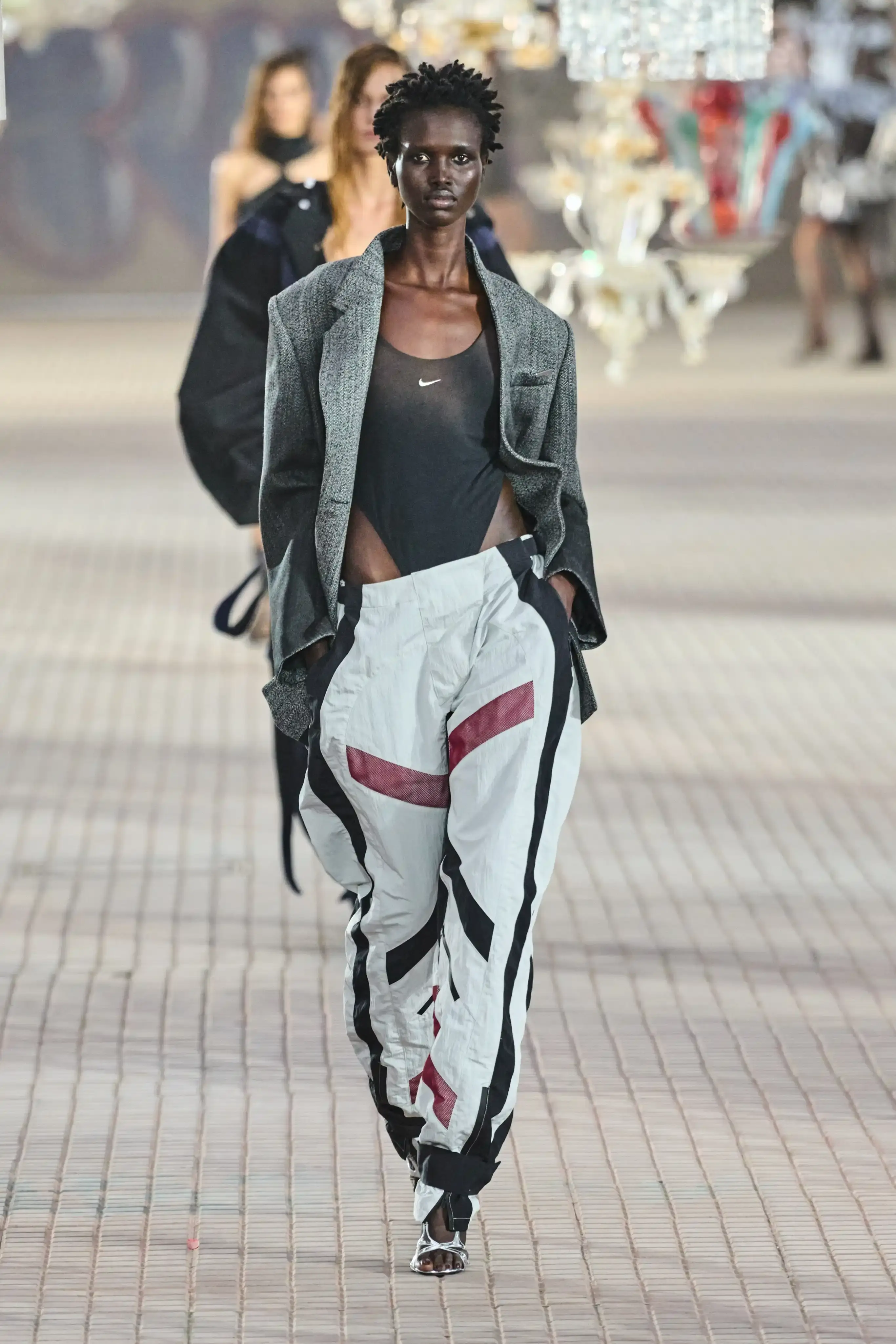 The Attico's Spring/Summer 2025 runway show at Milan Fashion Week features a surprising collaboration with Nike. Models walk on an industrial set adorned with colorful glass chandeliers, showcasing a collection that blends high fashion with sportswear elements. The runway presentation, titled "The Sound of Breaking Glass," explores themes of fragility and strength through contrasting designs. Sheer fabrics, form-fitting silhouettes, and embroideries are juxtaposed with nylon materials, bomber jackets, and cargo pants. The color palette is predominantly dusty and faded. Select Nike athletic wear pieces are integrated into the collection, symbolizing resilience and personal growth. Designers Gilda Ambrosio and Giorgia Tordini aim to empower women with this innovative fusion of luxury fashion and athletic functionality, challenging traditional runway norms (image 3 of 6)