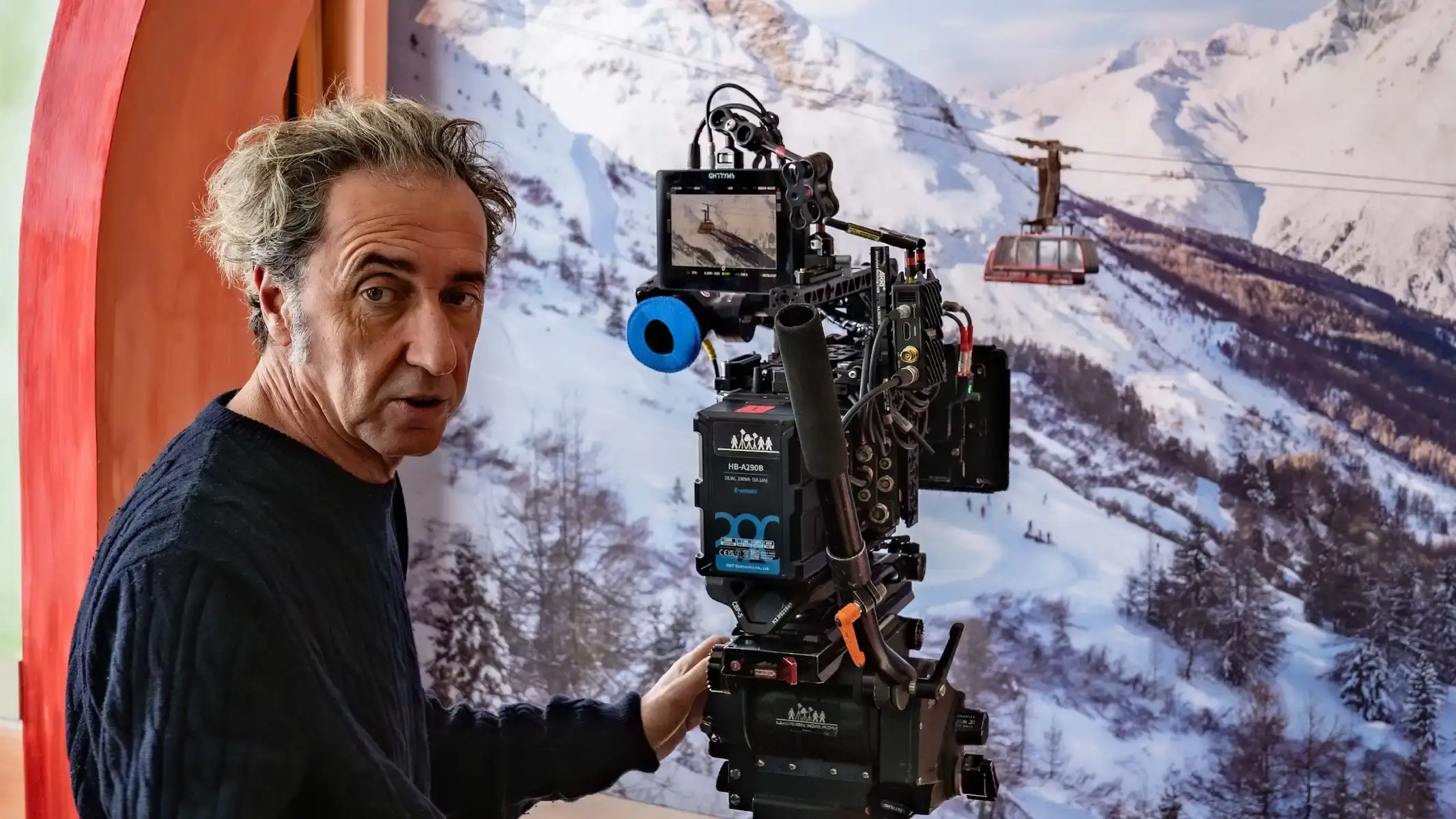 Paolo Sorrentino directing a winter-themed short film for Moncler Grenoble, showcasing ski wear and mountain gear in domestic settings with people performing winter sports activities indoors